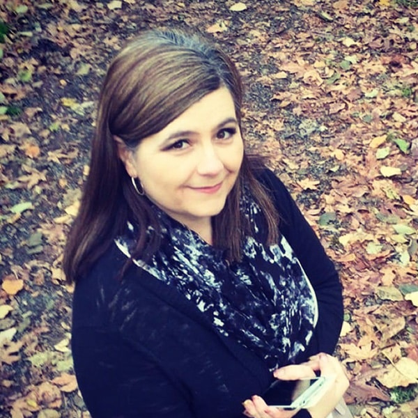 A person with brown hair stands outdoors on a path covered with fallen leaves, holding a smartphone and wearing a dark sweater and patterned scarf.