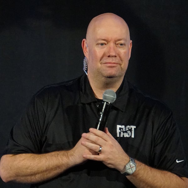 A person wearing a black shirt holds a microphone while standing against a dark background.